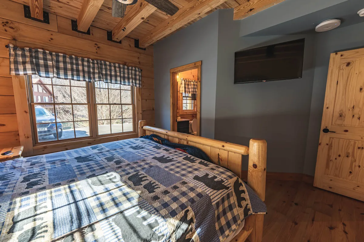 A bedroom with a wooden bed frame, plaid blanket, and large windows that let in ample natural light. The view from the windows suggests the surrounding mountains and forests, reinforcing the cabin's setting in the heart of the Smokies.