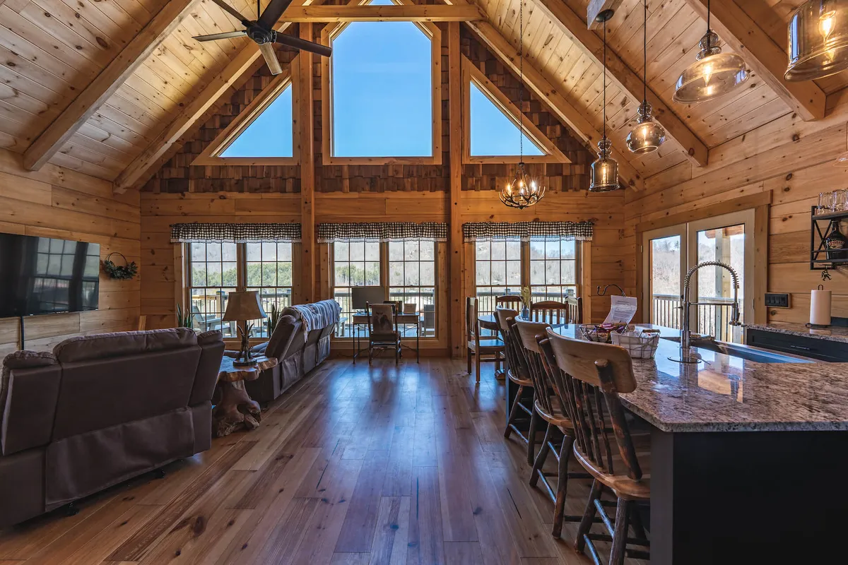 A bright, airy living-dining area with comfortable seating, a kitchen island, and large windows streaming in natural light. The rustic wooden beams and furnishings evoke a sense of cabin comfort and togetherness.