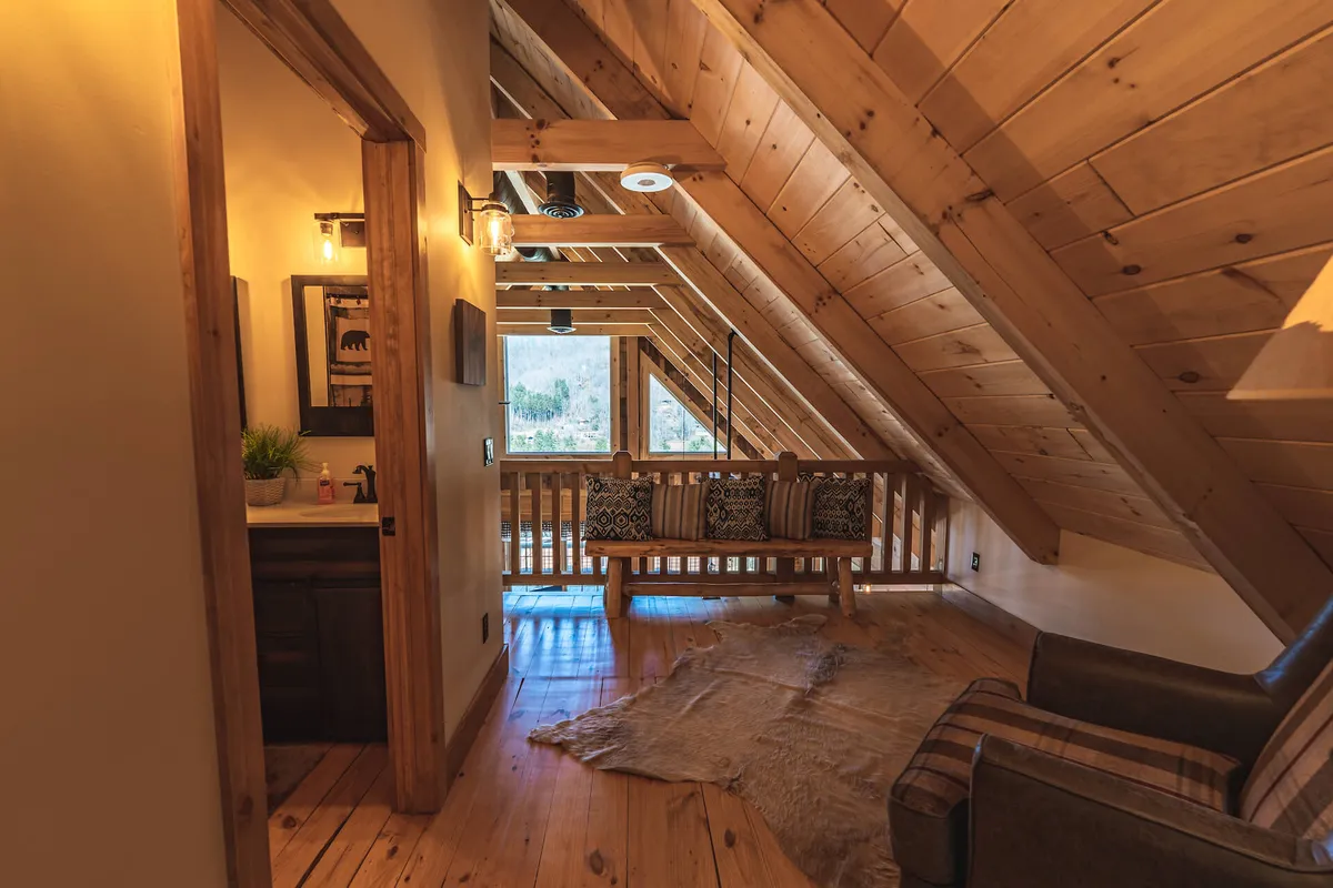 This image shows a cozy seating area with exposed wooden beams and a swing bench positioned by a window. The window offers a view outside, hinting at the beautiful natural surroundings of the cabin. The space invites relaxing, reading, and enjoying the scenery beyond the walls.