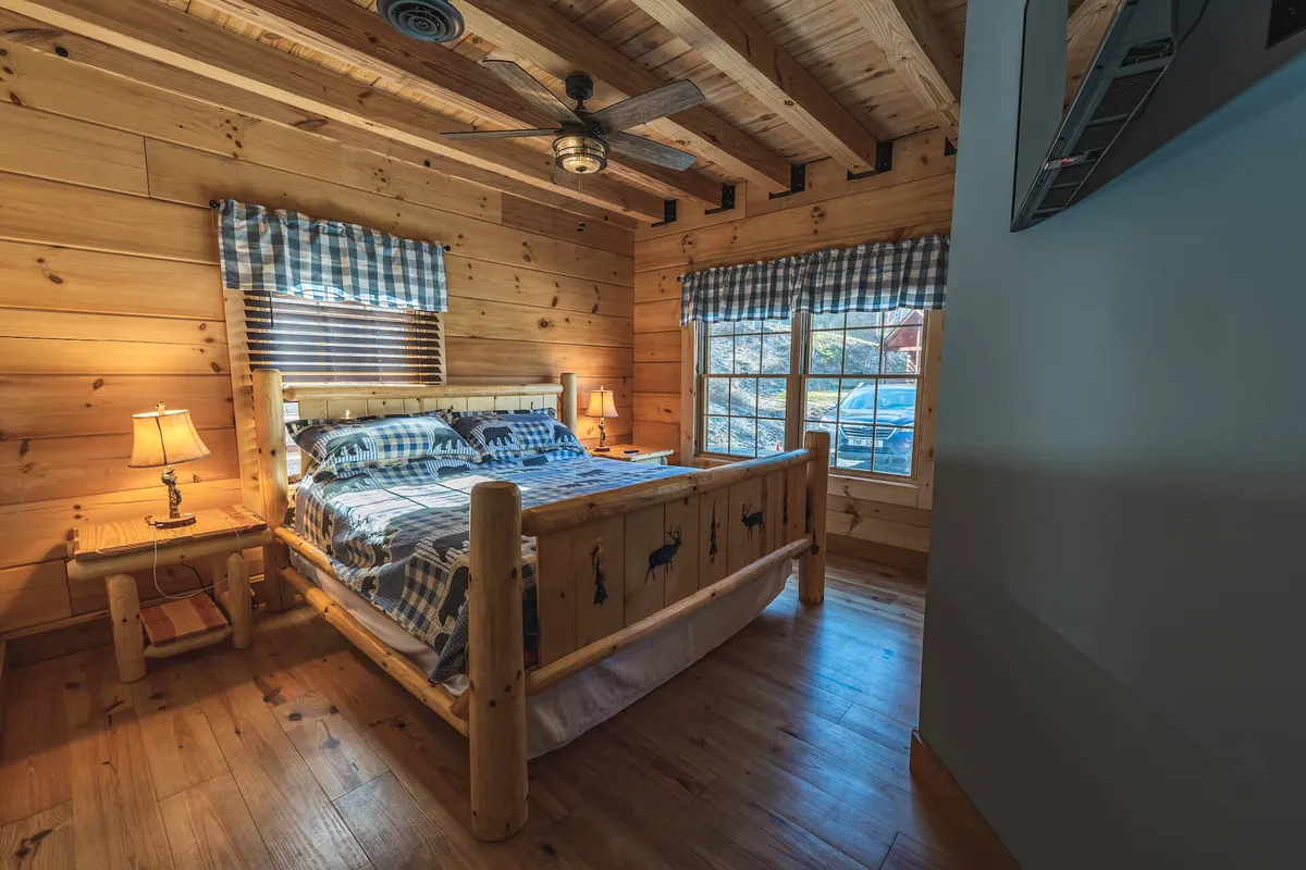 A cozy bedroom featuring a wooden bed with decorative pillows, warm lamps on bedside tables, and windows adorned with checkered curtains. Natural light gently filters through, creating a peaceful retreat ideal for relaxation, reading, or starting the day with a view of the Smokies.