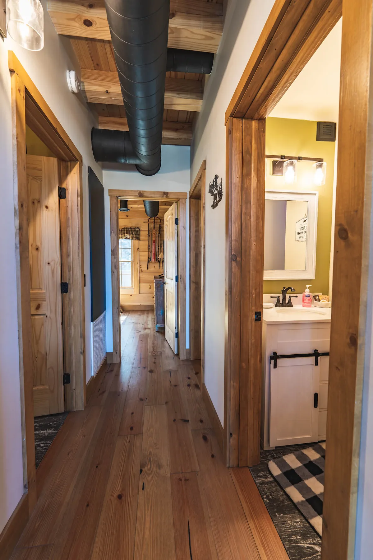 A corridor with warm wooden flooring, visible door frames, and a bathroom sink creates a sense of transition from the entryway into the heart of the cabin. The scene is cozy and practical, suggesting a seamless flow between utility spaces and the rest of the home.