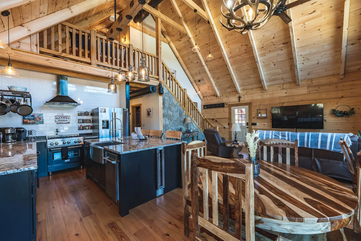 Offering a wider perspective, this image reveals the open layout connecting the kitchen island, dining table, staircase, and a striking stone wall. The scene captures the effortless flow between kitchen, dining, and living areas, underscoring the social and communal possibilities in the heart of the home.