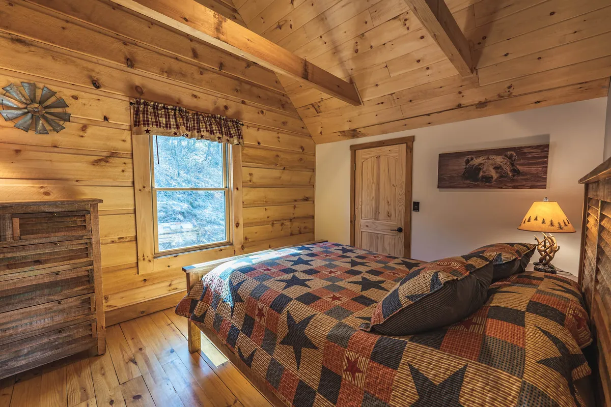 A welcoming bedroom with a wooden bed draped in a star-patterned quilt, a large window offering views, and a wooden dresser, all bathed in soft, natural light. The space exudes comfort and relaxation, ideal for couples or groups to unwind together.