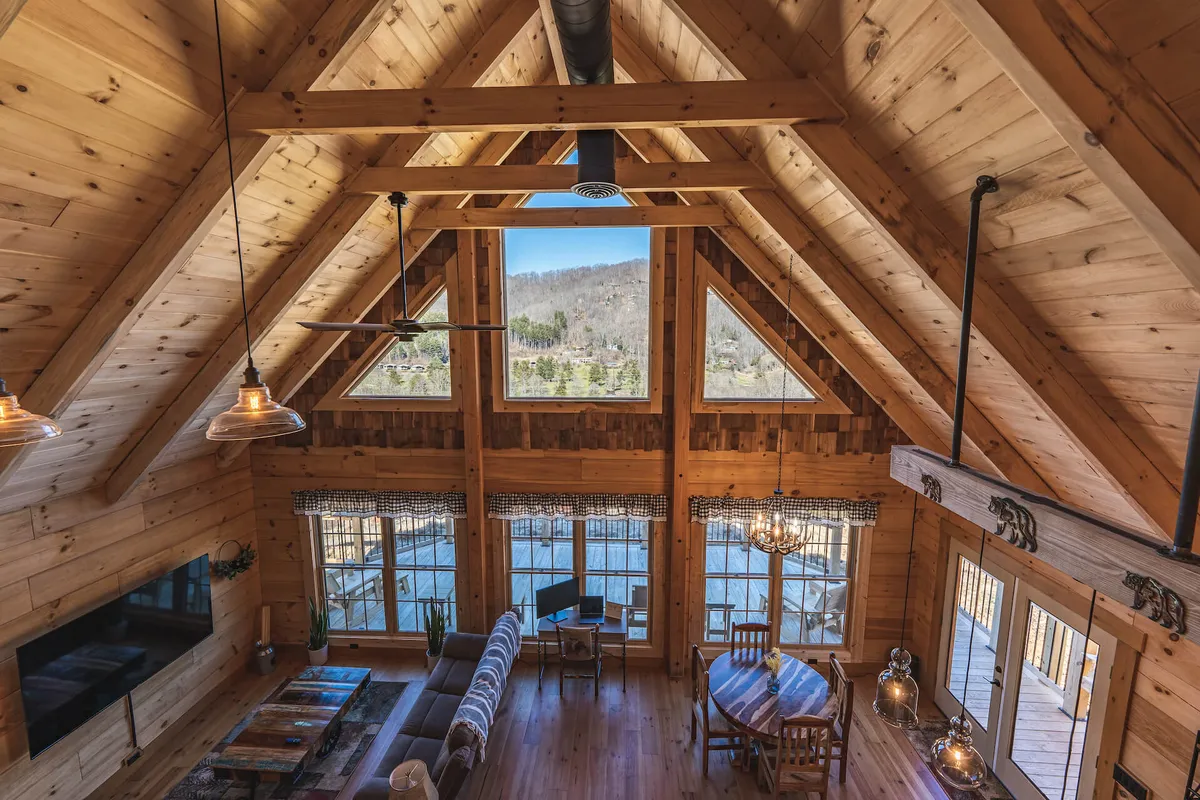 A spacious living area featuring large windows that perfectly frame sweeping views of the mountains. The room is appointed with comfortable seating, exposed wooden beams, a dining table, and warm lighting, creating an inviting communal space ideal for gatherings.