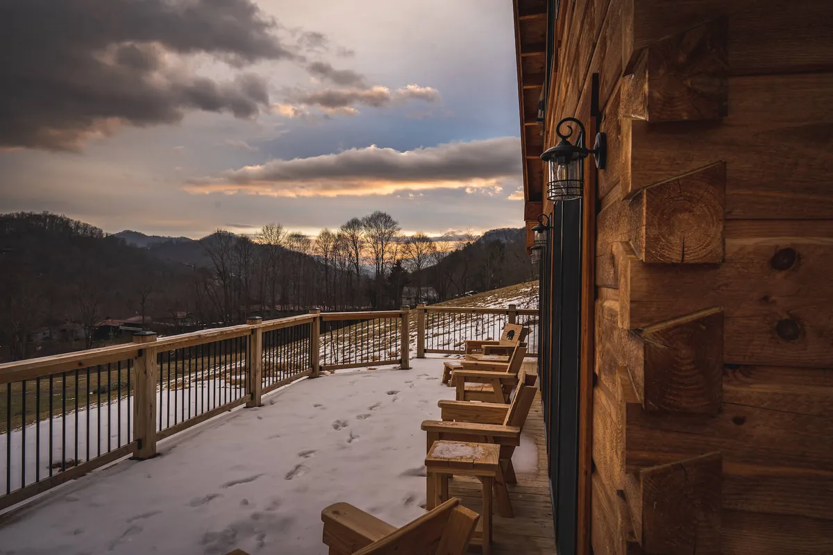 The hero image features the exterior of a luxurious log cabin framed by snow-dusted ground and wooden Adirondack chairs, all facing toward a sweeping view of the Smoky Mountains under a dramatic cloudy sky. The landscape evokes a sense of calm seclusion, inviting guests to relax, take in nature, and enjoy the changing sky—perfect for unwinding or stargazing.