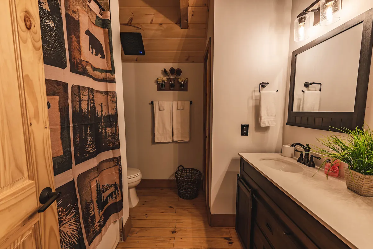 A warmly decorated bathroom with a wooden door, a bear-themed shower curtain, a bright vanity with a sink, and a touch of greenery from a potted plant. The space feels both inviting and practical, perfect for unwinding after a day of adventure.